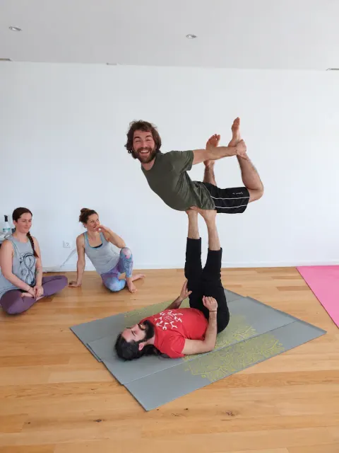 A smiling flyer holds bow pose above a partner lying on a yoga mat, while two people watch from the studio wall.