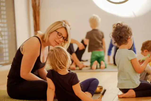 A woman sits beside a young child on the studio floor while other children and adults practise behind them.