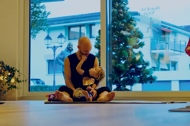 An adult and young child sit together on a yoga mat beside a Christmas tree in a window-lined studio.