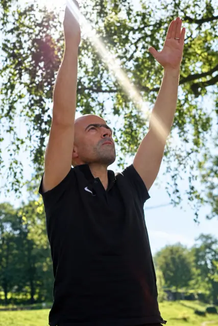 A man in a black shirt looks upward with both arms raised as sunlight filters through the leafy trees above him.