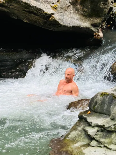A shirtless man sits in a rushing pool beneath a small waterfall, surrounded by smooth rocks and white water.