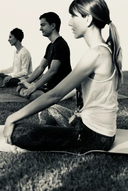 Three people sit cross-legged with hands resting on their knees during an outdoor meditation on the grass.