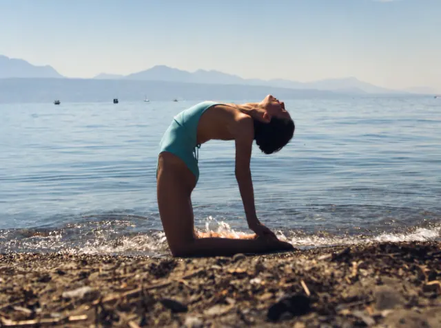 A woman holds camel pose on a pebbled lakeshore, with calm water and distant mountains behind her.