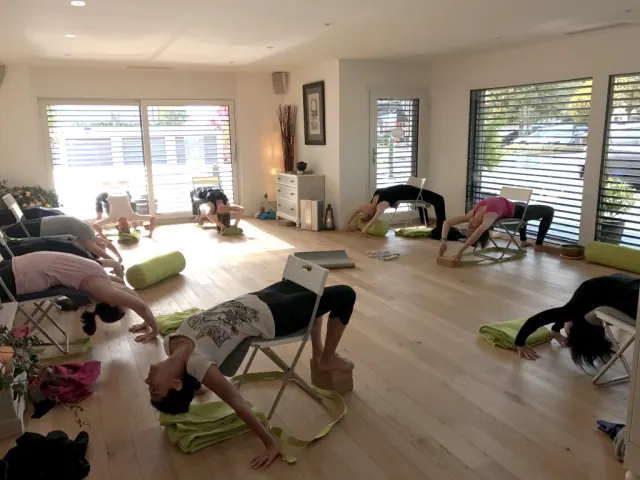 Participants practise supported yoga backbends using chairs and bolsters in a bright studio with large windows.