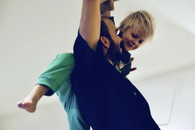 A smiling child rides on a man's shoulders while he raises his arms during an indoor movement session.