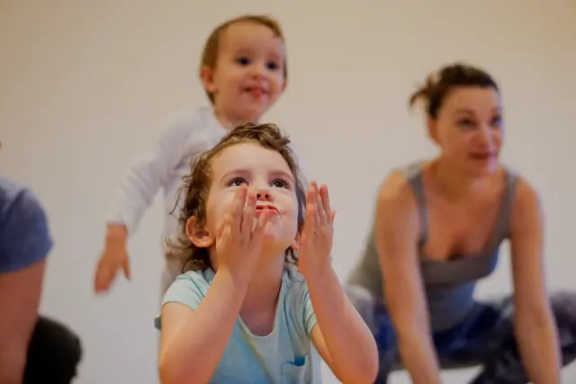 A young child looks upward with both hands raised beside her face, with another child and an adult behind.