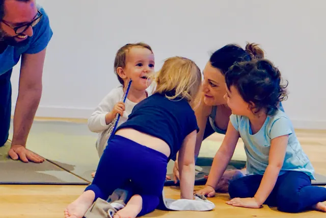 Children and adults gather closely on yoga mats, smiling and interacting during a playful studio session.
