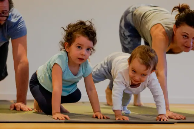 Two young children move on their hands and knees across yoga mats while adults join them in the studio.