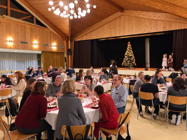 Guests share a festive meal around tables in a wooden hall, with a Christmas tree and performers on the stage.