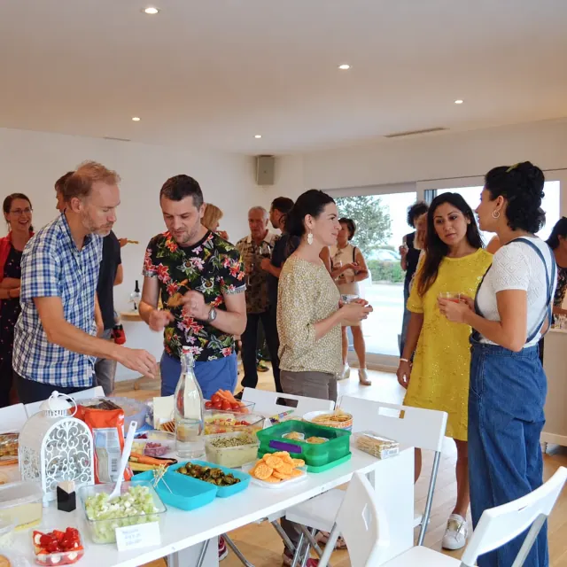 Adults chat around a table of colourful shared food in a bright room during a community gathering.
