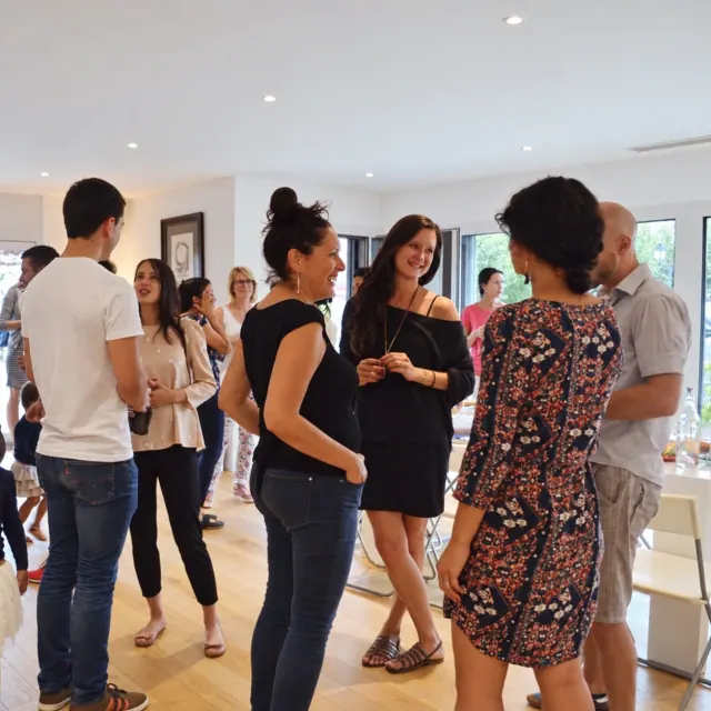 People stand chatting together at a community gathering in a bright yoga studio with wooden flooring and framed pictures.