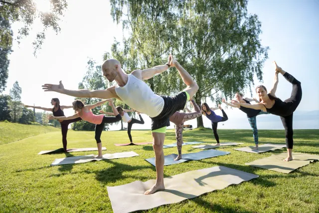 A teacher and group balance in dancer pose on a lakeside lawn, reaching forward while holding one foot behind.