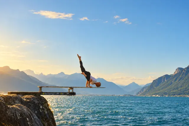 A man balances on his hands on a diving board above a blue lake, with one leg raised and sunlight on the water.