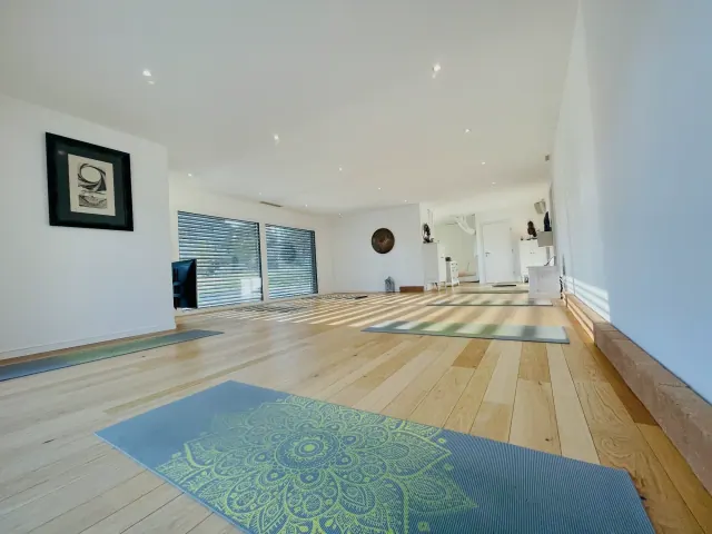 Yoga mats rest on the wooden floor of an empty white-walled studio, with sunlight streaming through the windows.