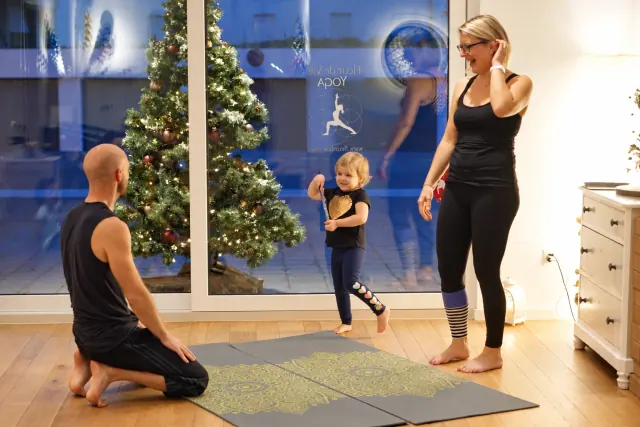 A child stands beside a woman during a family yoga session, with a kneeling teacher and a Christmas tree nearby.