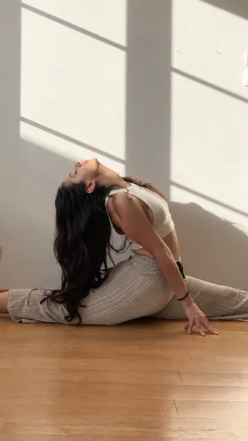 A woman arches backward in a front split on a wooden floor, with strong window shadows across the wall.