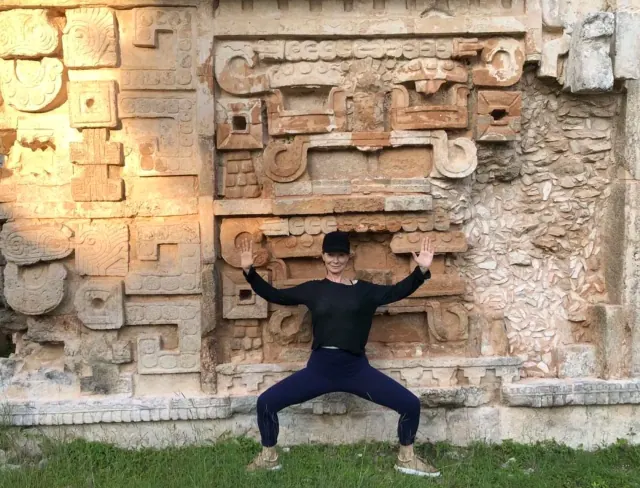 A woman holds a wide-legged Goddess pose with her arms raised in front of an intricately carved stone wall.