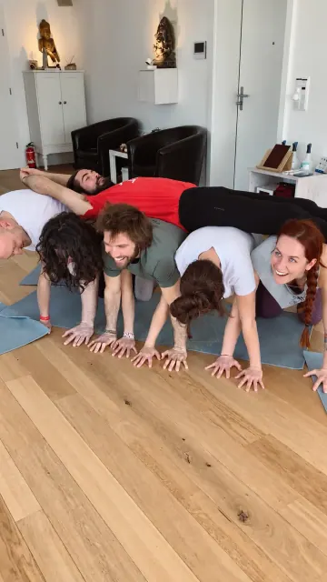 A man lies horizontally across the backs of five kneeling participants during a playful group acroyoga balance indoors.