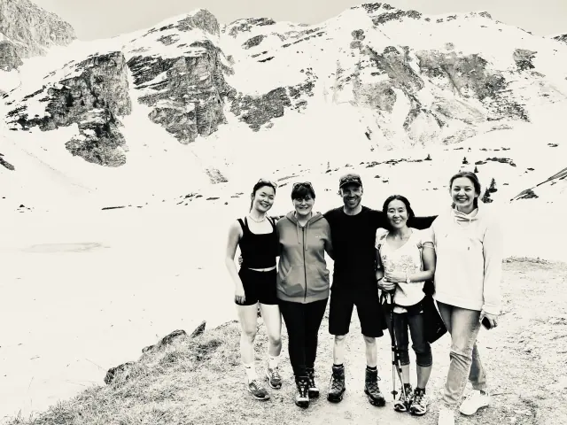 Five people stand together beside an alpine lake and snowy mountains in a black-and-white group portrait.