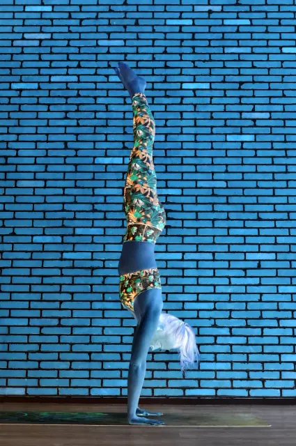 A woman in patterned leggings holds a handstand against a brick wall, shown in a stylised blue-toned composition.