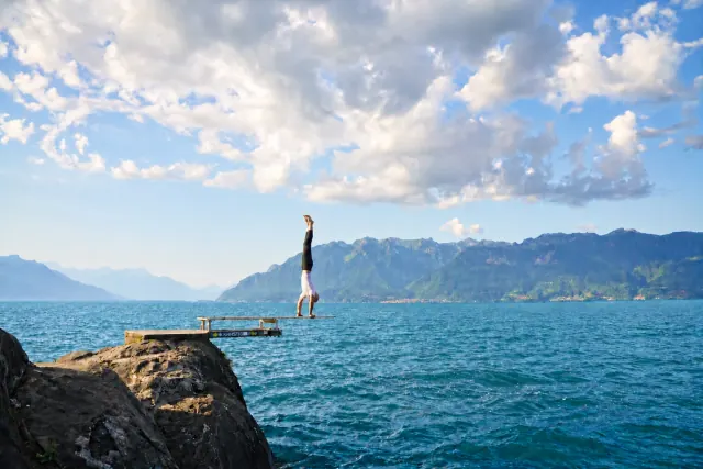 A man holds a handstand on a narrow board above a blue lake, with rocky shores, mountains and clouds beyond.