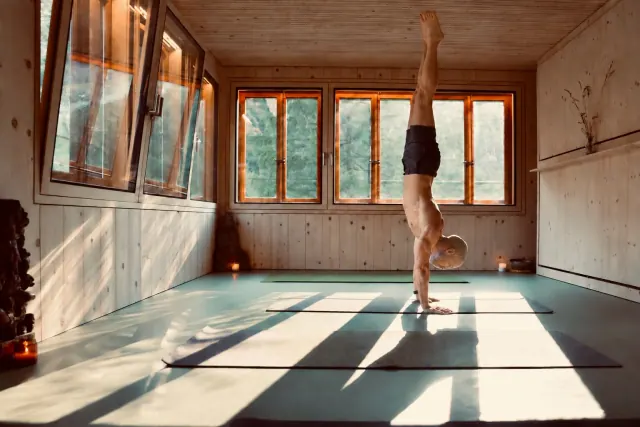 A man holds a straight handstand in a wooden yoga room, with sunlight from the windows casting long shadows on the floor.