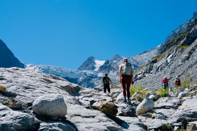 A group of hikers follows a rocky alpine trail toward a glacier, surrounded by steep mountains under blue skies.