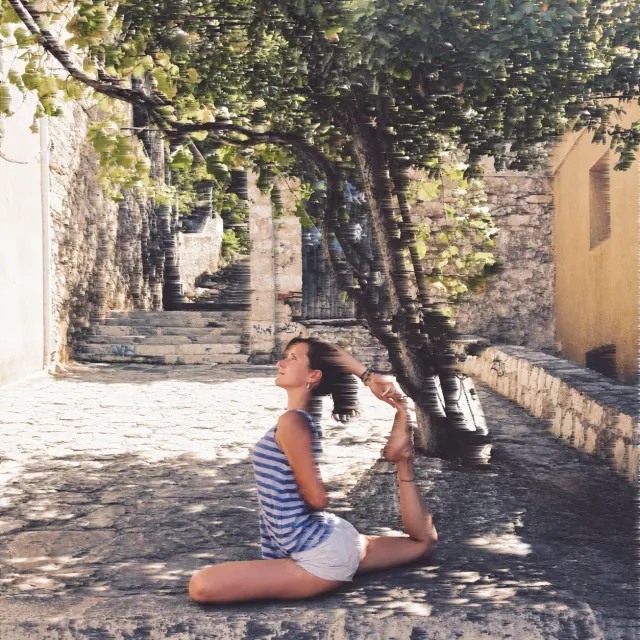 A woman holds king pigeon pose on a cobbled lane beneath leafy trees, lifting her back foot toward her head.