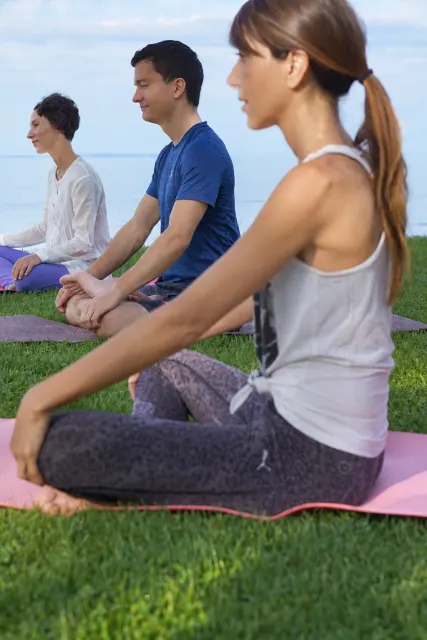 Three people sit cross-legged in a row on yoga mats beside a lake, with their hands resting on their knees.