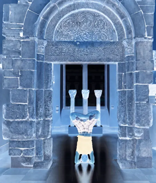 A colour-negative photograph of a person holding a lotus headstand beneath an ornate carved stone arch.