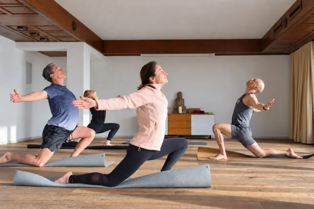 Three people practise low lunges with their arms open in a sunlit yoga room with wooden ceiling beams.