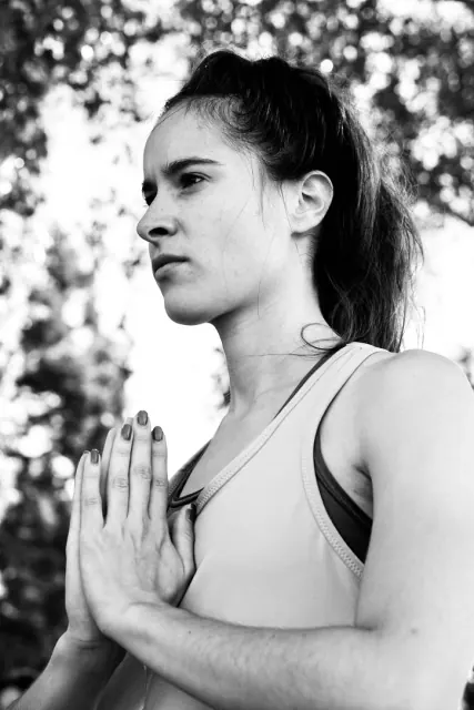 Black-and-white portrait of a woman holding her palms together at her chest, with softly blurred trees behind her.