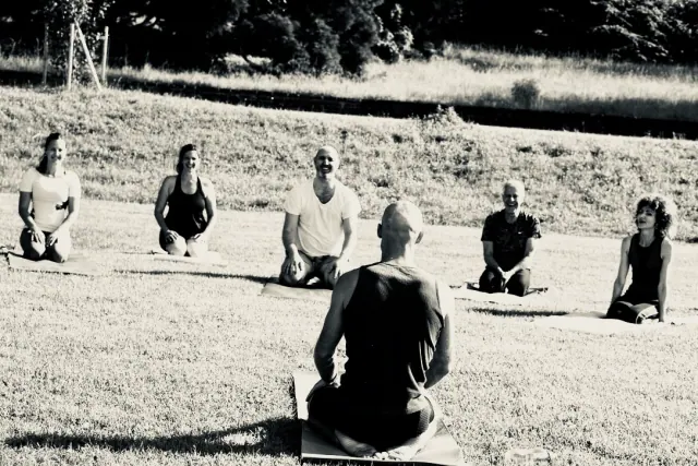 A black-and-white photograph of a teacher facing a seated yoga group arranged in a circle on the grass.