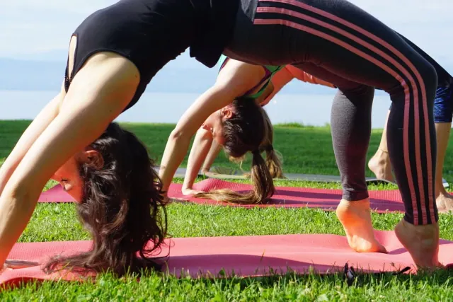 Women practise wheel pose on pink mats laid on the grass, with the blue lake visible beyond them.
