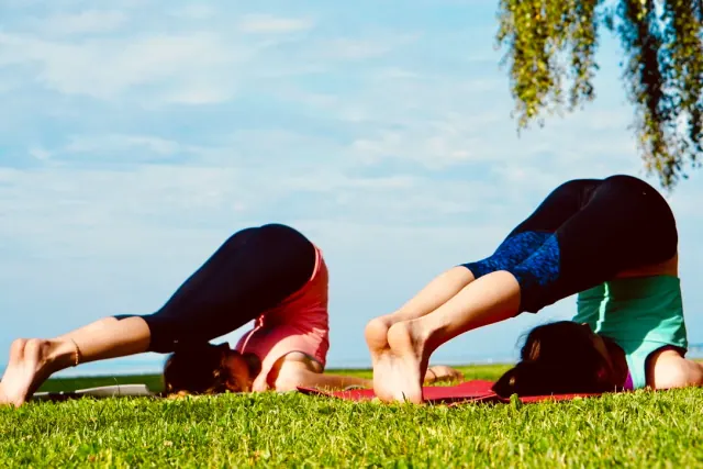 Two people practise plow pose on the grass beside a lake, with their legs folded overhead under a bright blue sky.