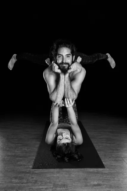 Black-and-white portrait of two partners holding an acroyoga balance, with one lying on a mat and supporting the other.