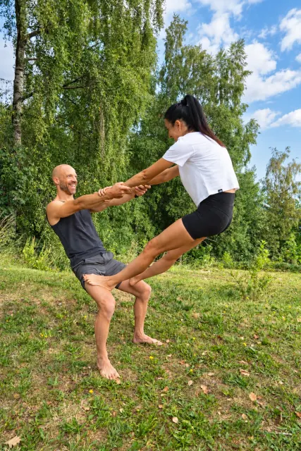 A smiling man supports a partner's feet on his thighs as they hold hands in a counterbalance beneath leafy trees.