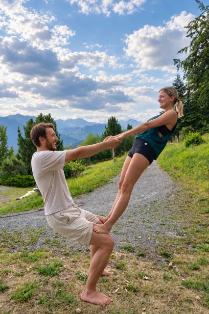Two smiling partners hold hands in a counterbalance on a grassy mountain path beneath a cloud-filled sky.