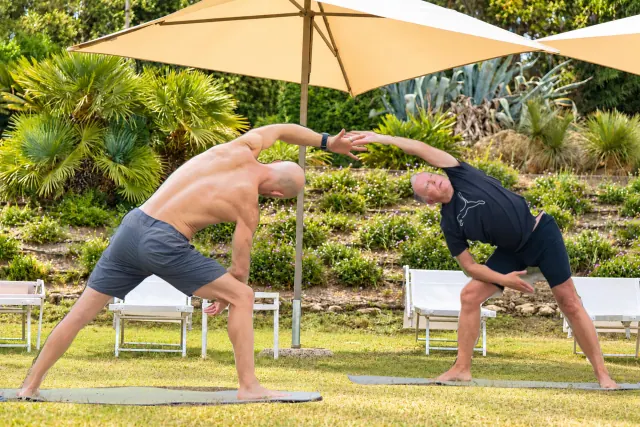 Two men lean toward each other in standing side stretches, joining their raised hands in a sunny garden.