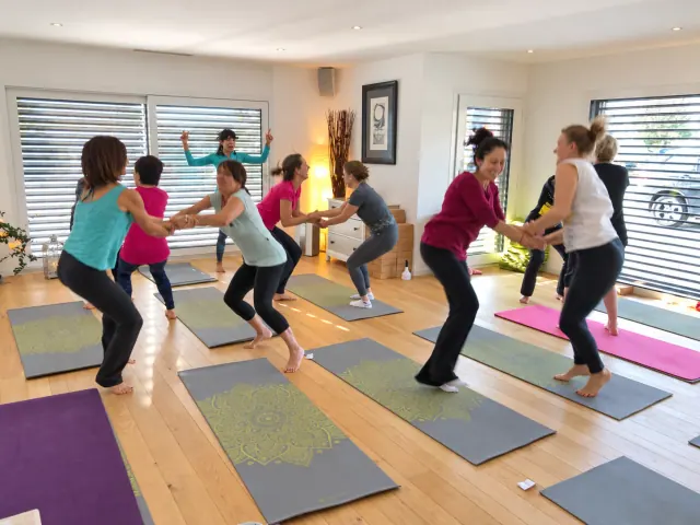 Pairs of women hold hands and bend their knees together on yoga mats in a bright, wood-floored studio.