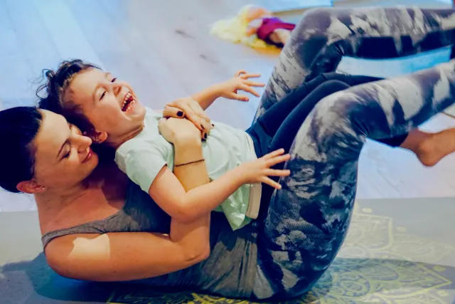 A woman cradles a laughing child while lying back with her knees raised during playful floor yoga.