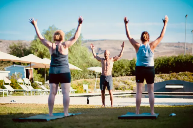 A man faces two participants as they raise their arms on yoga mats in a sunny garden with distant hills.