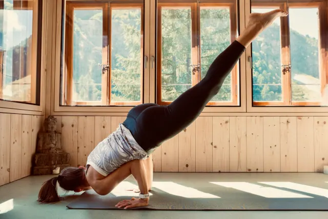A woman balances on bent arms with her legs extending upward in a sunlit, wood-panelled yoga room.