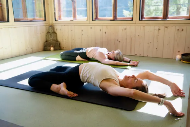 Two women recline with their knees folded and arms overhead on yoga mats in a sunlit, wood-panelled room.