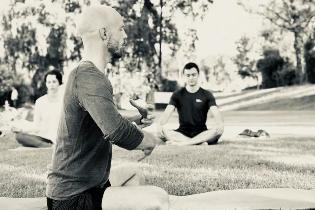 Black-and-white view of a seated yoga teacher gesturing with his hands while participants meditate on mats in a park.