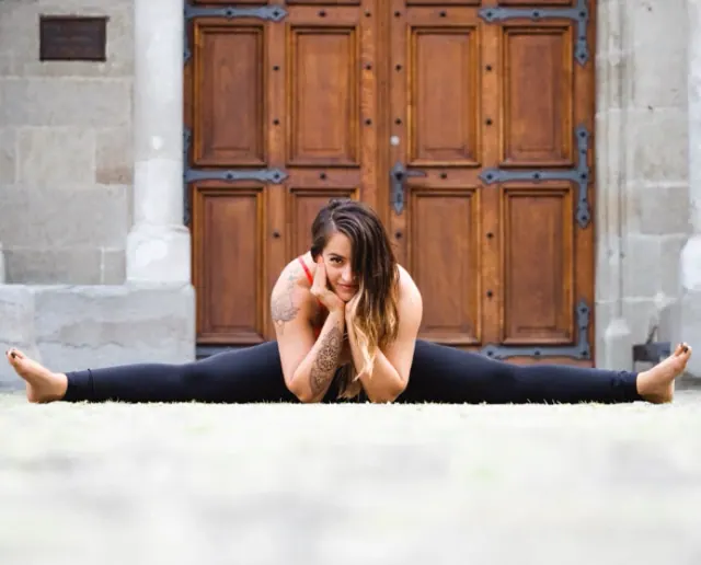 A woman rests her chin in her hands while sitting in a wide-legged split in front of a large carved wooden door.