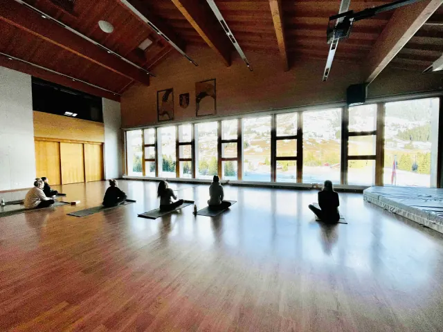 Seated participants face mountain-view windows in a spacious wooden hall, with daylight reflected across the floor.