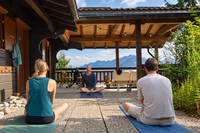 Three people sit cross-legged on yoga mats beneath a covered wooden terrace overlooking the mountains.