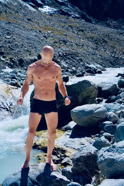 A man in dark swimming shorts stands on a rock beside a fast-flowing alpine river, looking down at his footing.