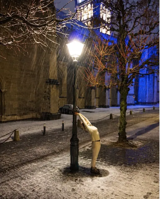 A woman stretches into a standing backbend beside a glowing streetlamp in a snowy square at night, near a stone church.